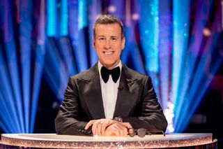 Anton Du Beke sitting at the judges' desk in the BBC Strictly studio. He is wearing a tuxedo and a black bow tie, and his glasses are on the desk in front of him. The room behind him is lit by bright blue and purple floor lights.