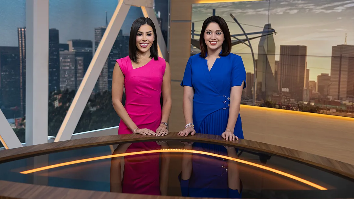 Two female anchors at a desk with city skyline in background