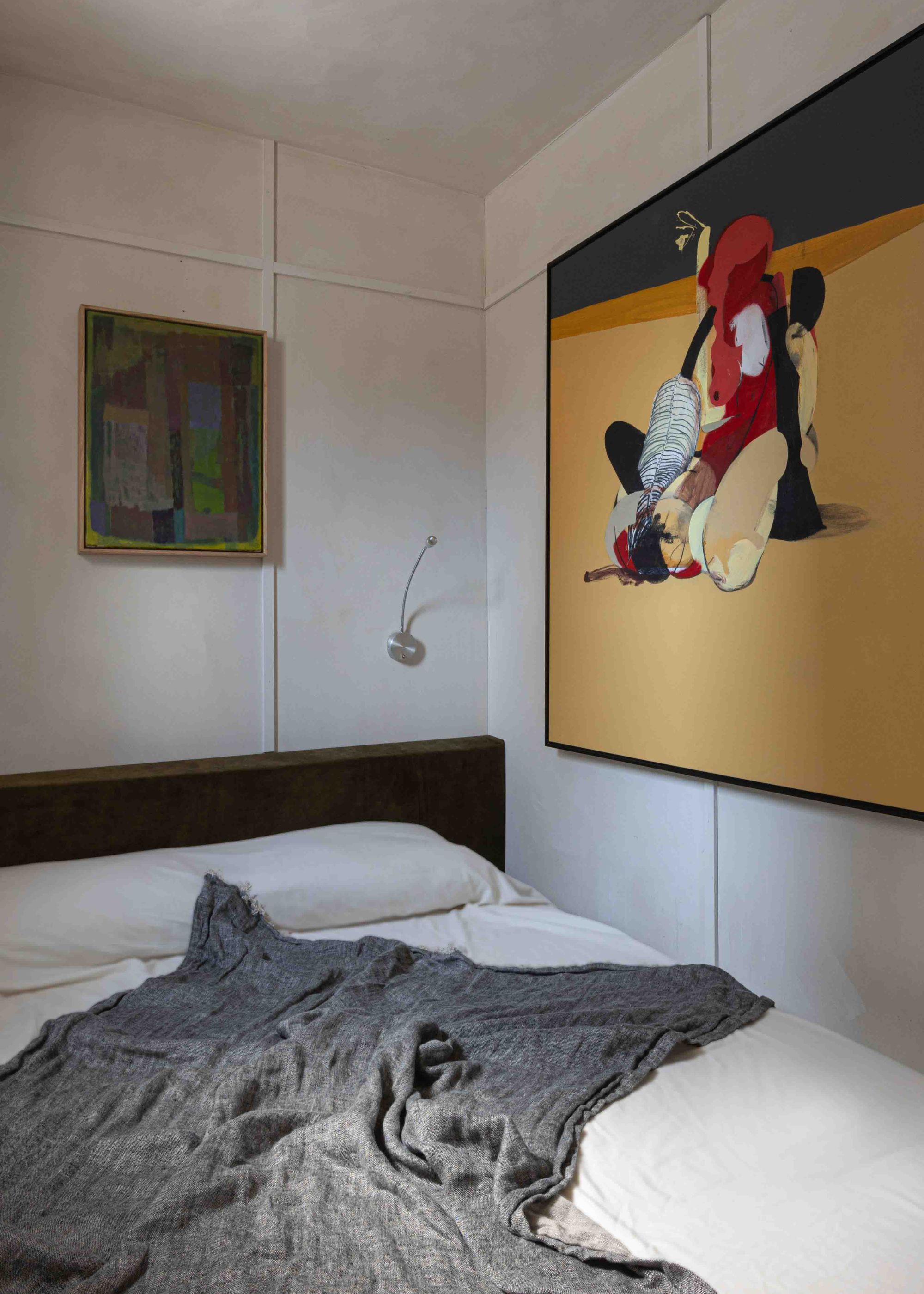 A bedroom with two large art pieces, a bed by the wall, white bedding and a gray throw