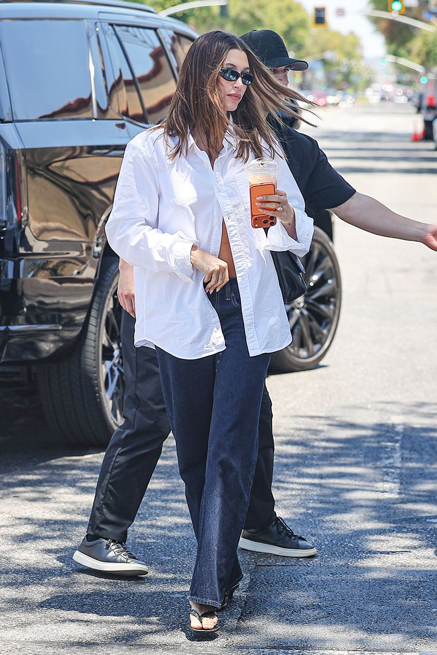 Hailey Bieber wearing a white button-down shirt with low-rise dark-wash jeans, kitten-heel thong sandals, and The Row shoulder flap bag