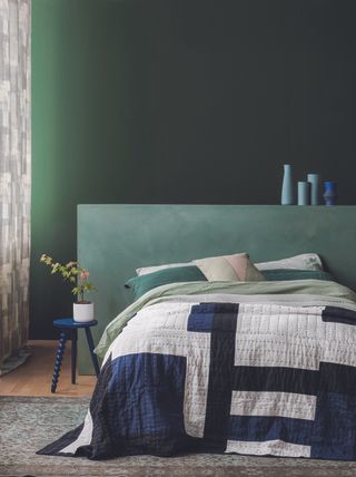 A green themed bedroom with green walls, a green oversized headboard and a blue and white quilt