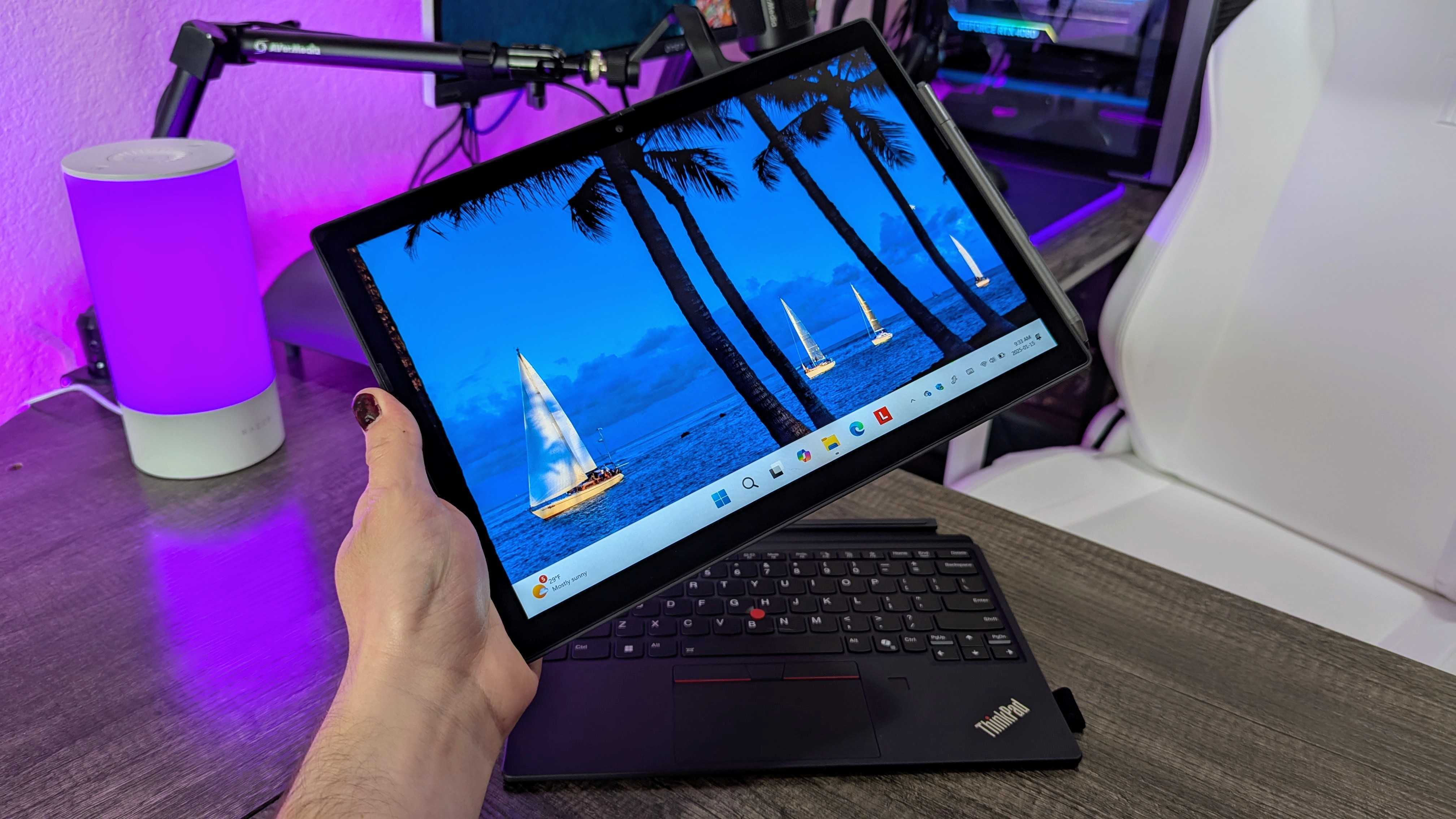 The Lenovo ThinkPad X12 Detachable (Gen 2) held in the hand with the stylus attached, with the detached keyboard lying on a desk in the background.