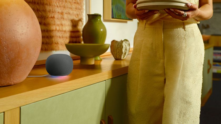 The Google Home Speaker in black, sitting on a countertop while a model walks by.