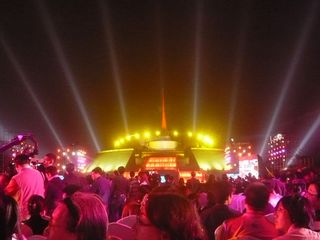 Crowds of people at an opening ceremony. The building is lit in yellow and spotlights fan out into the sky
