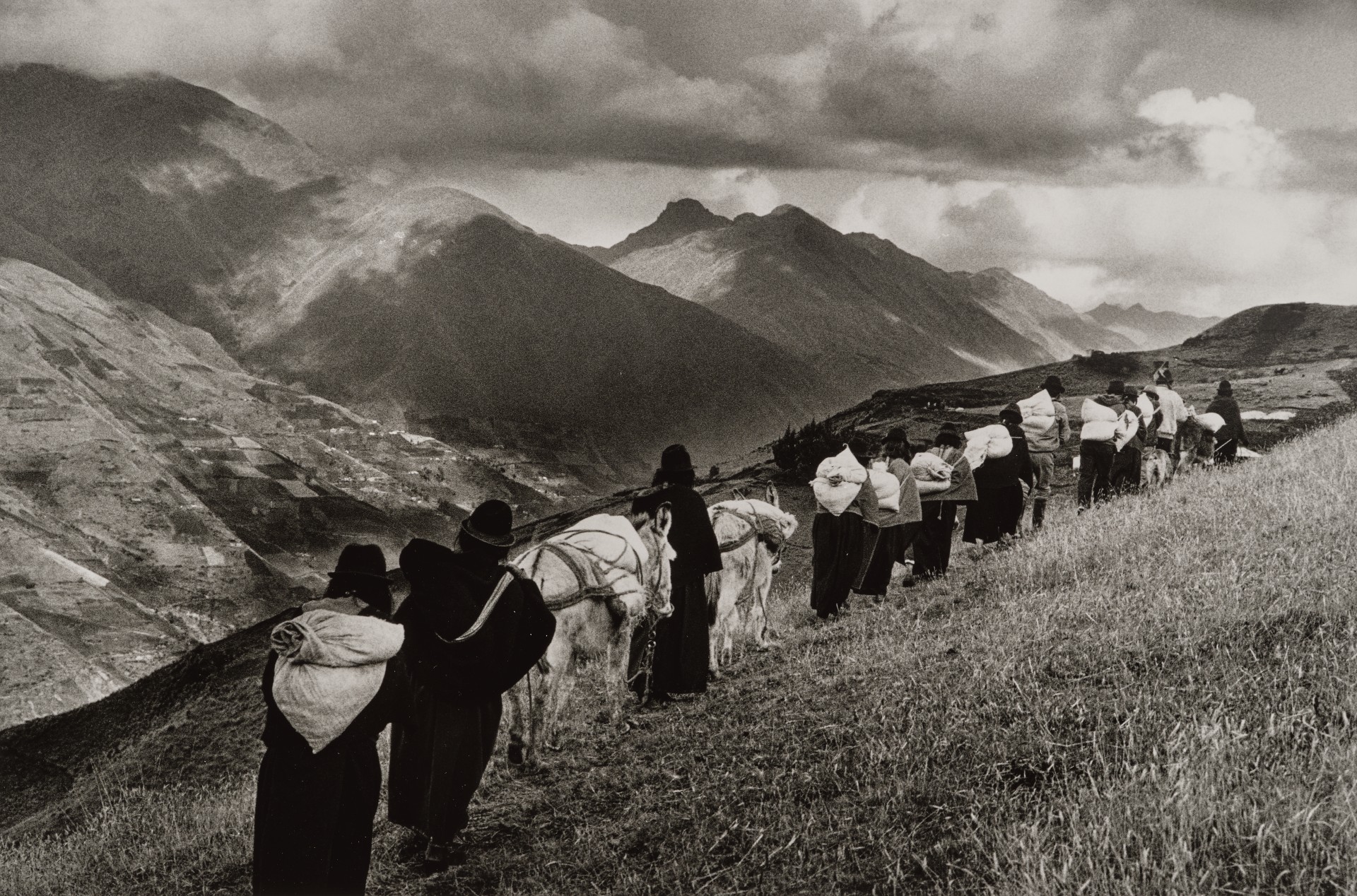 A black-and-white photograph of a long line of women and white-loaded donkeys walking in single file along a grassy mountain ridge, with dramatic peaks and storm clouds filling the background.