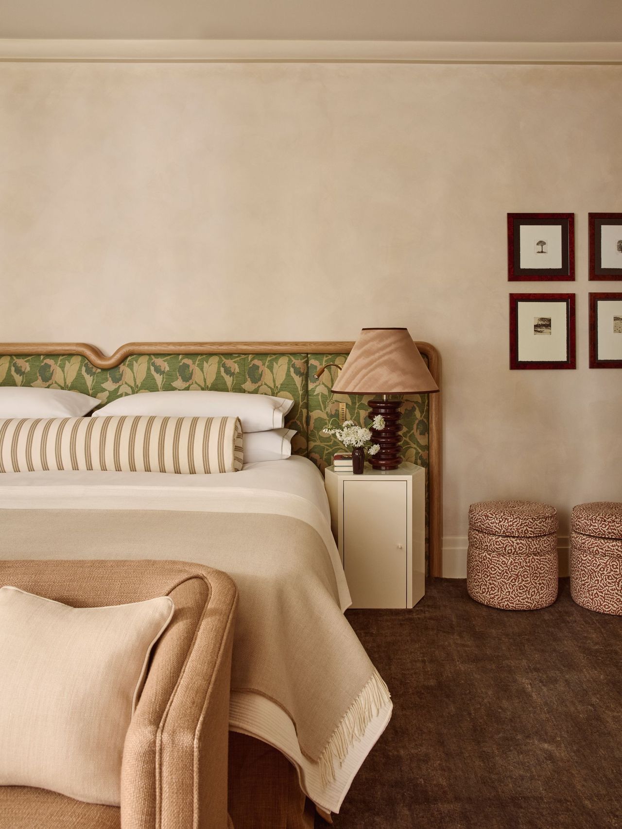 a bedroom with a green floral headboard, a striped bolster pillow, a tan blanket, and white pillows. A brown rug rests beneath the bed and two ottomans sit to the right
