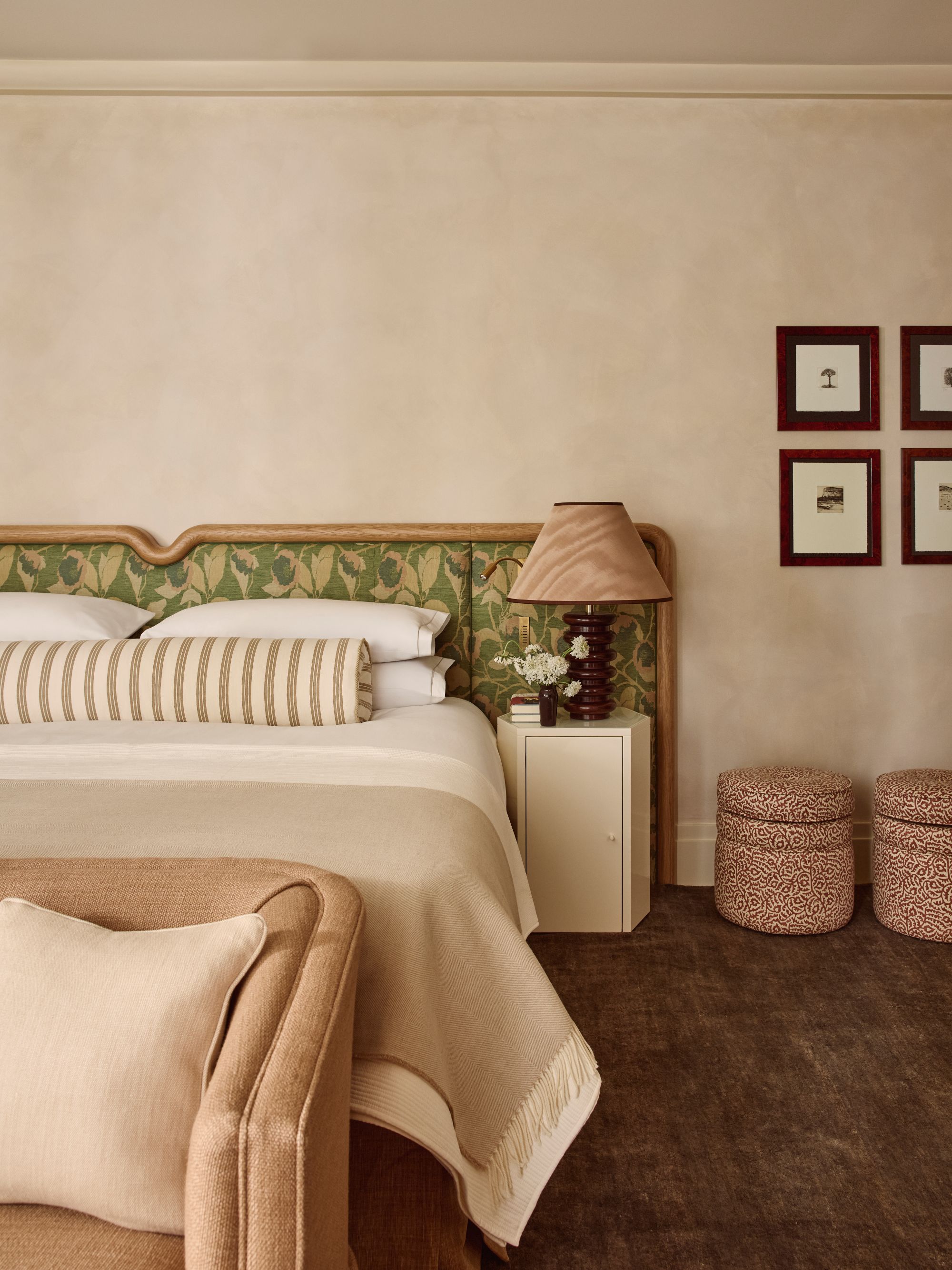 a bedroom with a green floral headboard, a striped bolster pillow, a tan blanket, and white pillows. A brown rug rests beneath the bed and two ottomans sit to the right