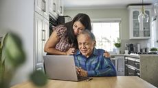An adult daughter hugs her aging father while helping him with something on a computer at the kitchen table.