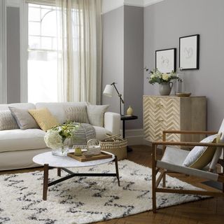 grey living room with wall-mounted picture frames above a wooden chest drawer and a cream-colored sofa with pillows and a circle marble coffee table on top of a wool rug