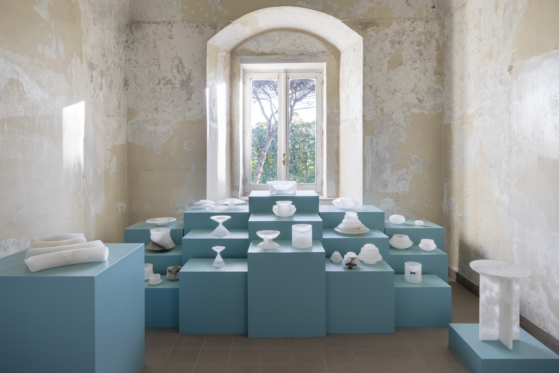 A design installation of matte glassware sits atop blue-tinted rectangular podiums in a historical building with distressed cement walls and an arched window overlooking greenery.