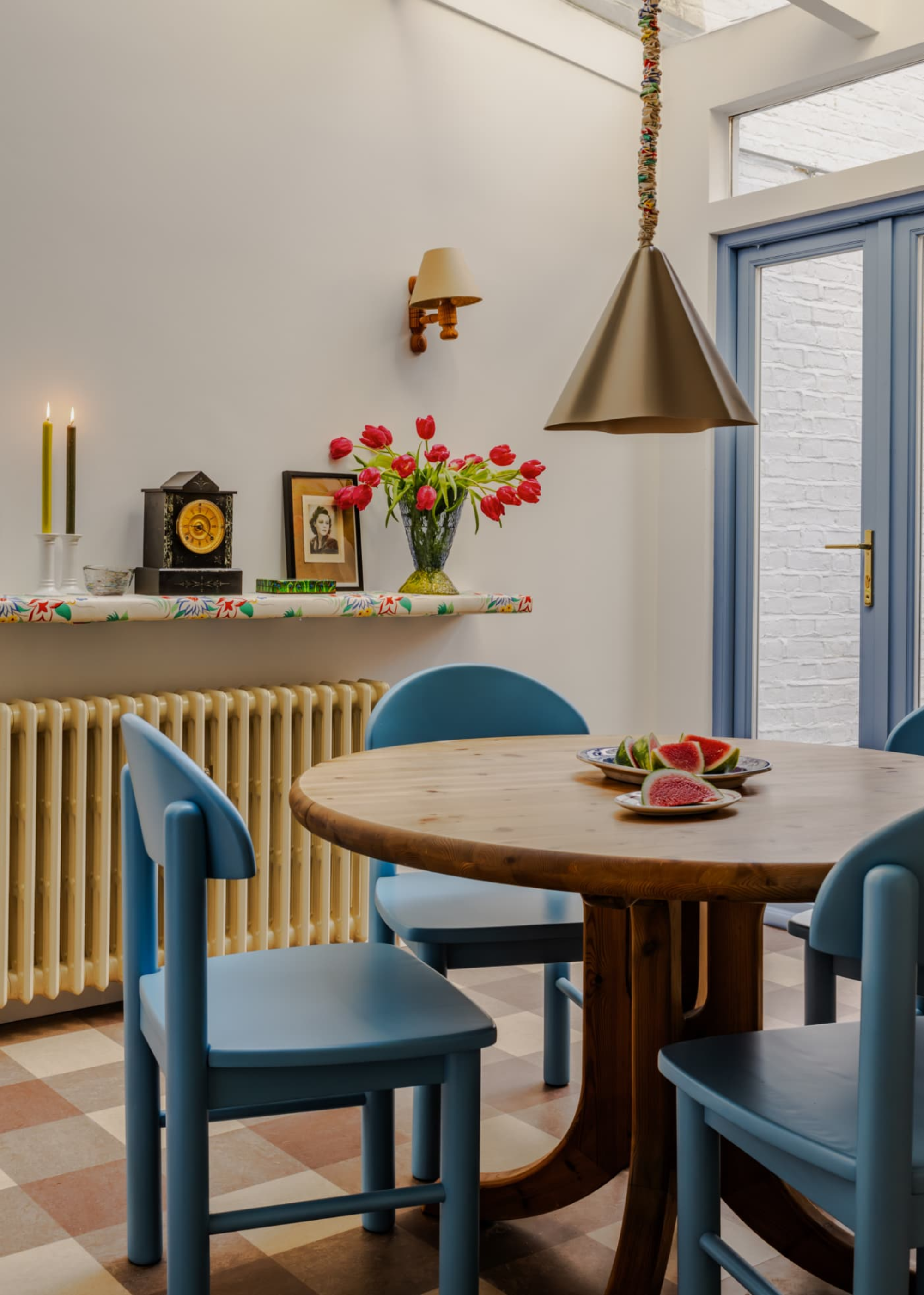 modern dining room with round table, blue dining chairs, a pendant light overhead, yellow radiator, and floral floating shelf with clock, candles, picture, and vase of flowers