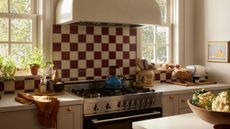 A close-up of a cozy cottage kitchen with neutral cabinets in front of two windows with a cooker in the middle with burgundy and cream checkerboard backsplash tiles. 