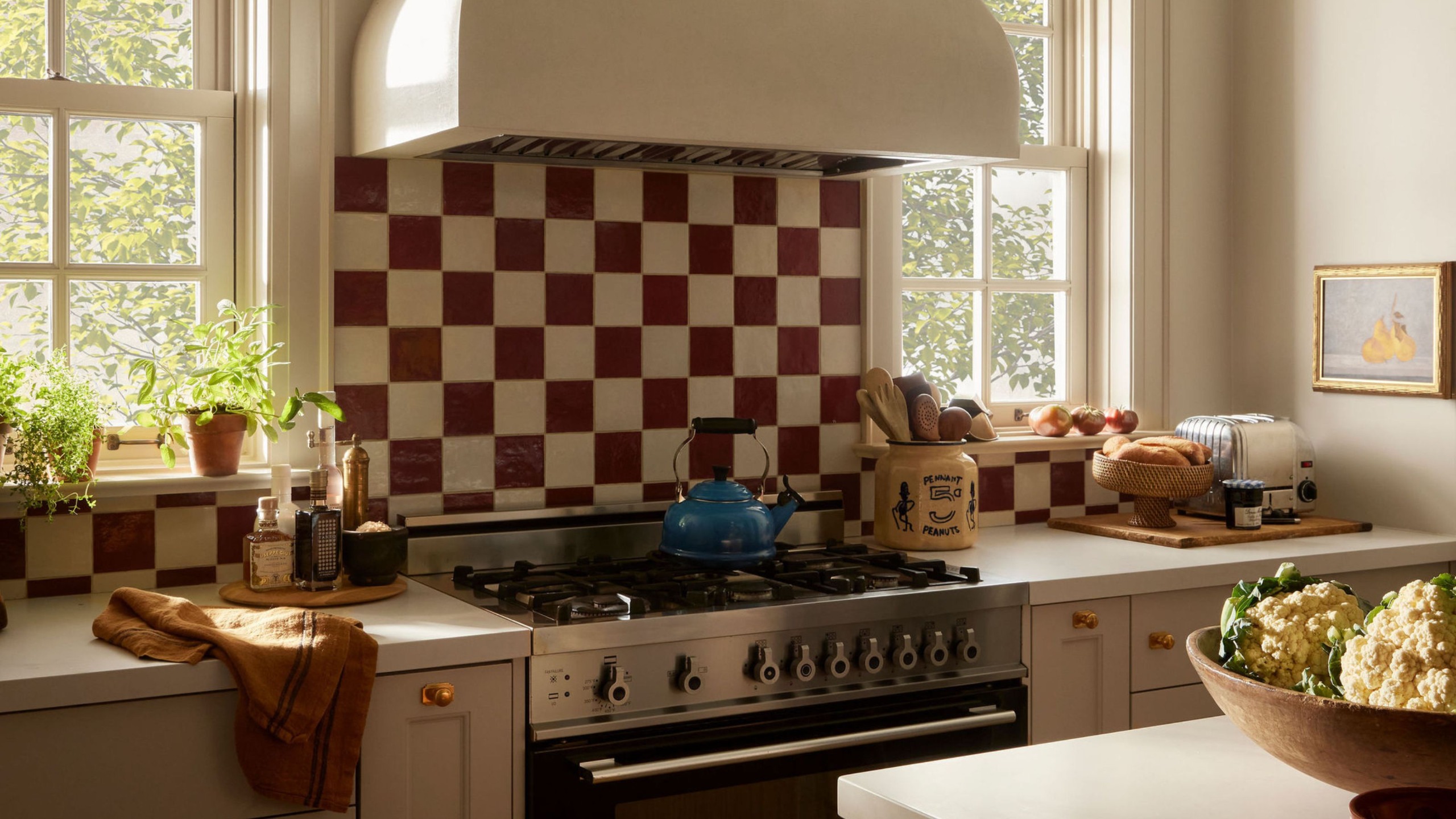 A close-up of a cozy cottage kitchen with neutral cabinets in front of two windows with a cooker in the middle with burgundy and cream checkerboard backsplash tiles. 