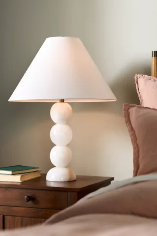 A white, crystal bobbin beside a table lamp with a white shade on a dark wooden bedside table. There is the side of a bed with a muted pink bedding in view