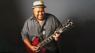 Larry McCray smiles as he plays his cherry red Guild Polara. He wears a black-and-gray striped shirt and grey pork pie hat