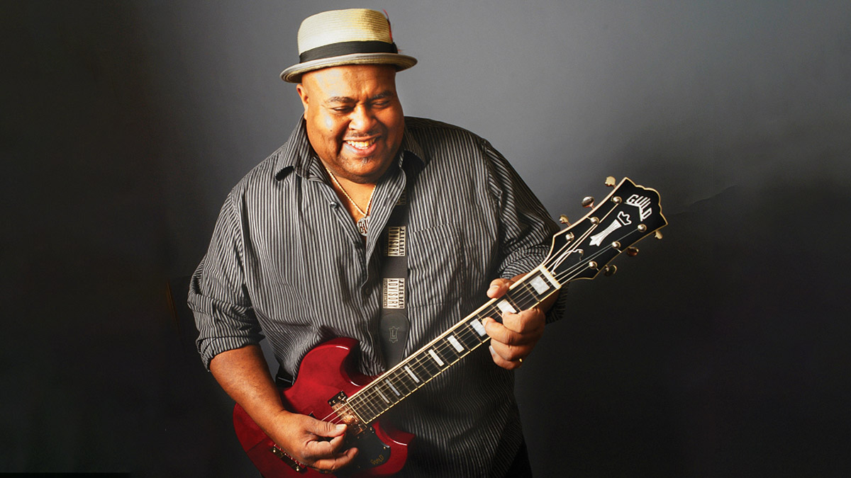 Larry McCray smiles as he plays his cherry red Guild Polara. He wears a black-and-gray striped shirt and grey pork pie hat