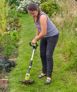 woman using a cordless weed eater/strimmer to cut grass path