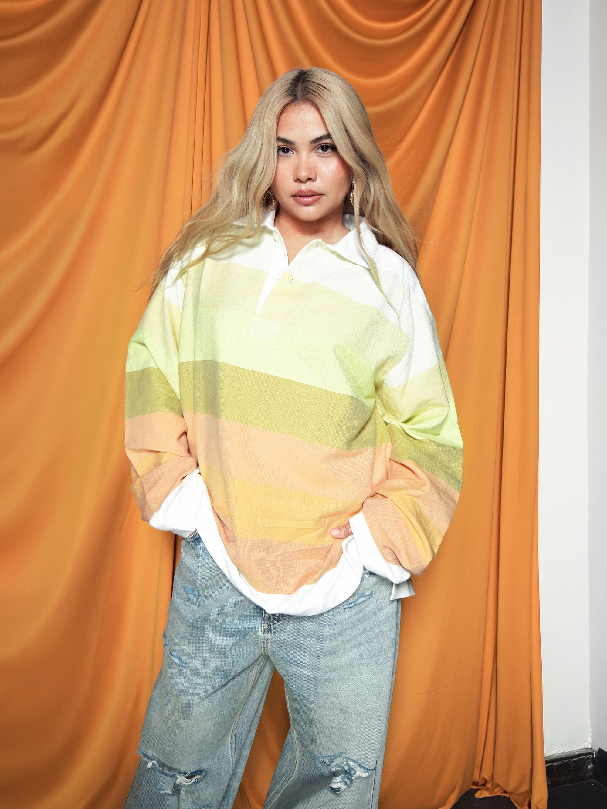 Hayley Kiyoko is standing with her hands in her pockets in front of an orange fabric backdrop, wearing an oversize collared long-sleeve shirt with blue jeans.