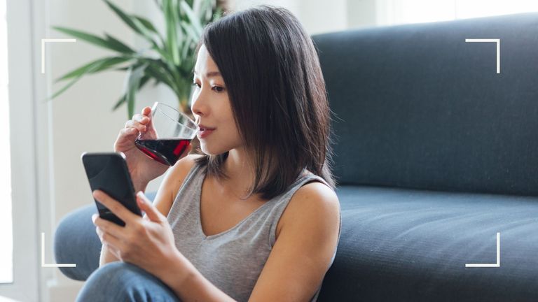 Woman drinking wine at home, researching how long do hangovers last on phone in front of a blue sofa