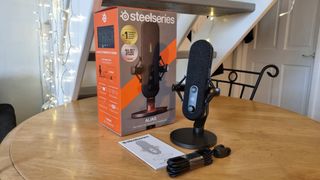 The SteelSeries Alias microphone, standing on a desk next to its box