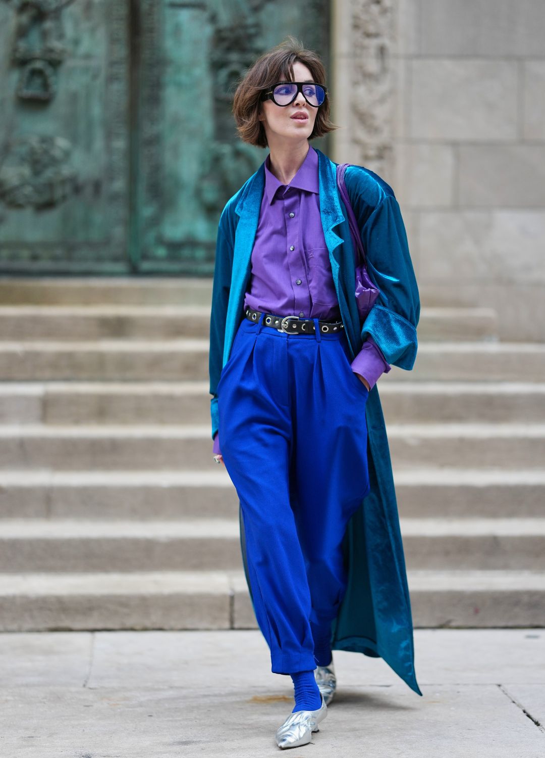 A guest wears a short tousled brown bob with fringe, oversized square sunglasses with blue-tinted lenses and dark frames, a silver ring on the right hand, a small shiny purple shoulder bag with a short strap, a teal velvet long duster coat over a purple button-down shirt, a black leather grommet belt cinching high-waisted cobalt blue pleated trousers with tapered cuffs, bright blue ribbed socks, silver metallic pointed-toe flats, outside Prabal Gurung, during New York Fashion Week, on September 13, 2025 in New York, New York