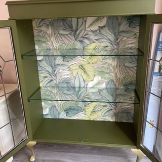 Restored and painted empty cabinet with glass shelves and doors with floral wallpaper backing on wooden floor