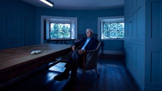 John le Carré (David Cornwell) sitting in a chair with his legs crossed in a room glowing with blue light.