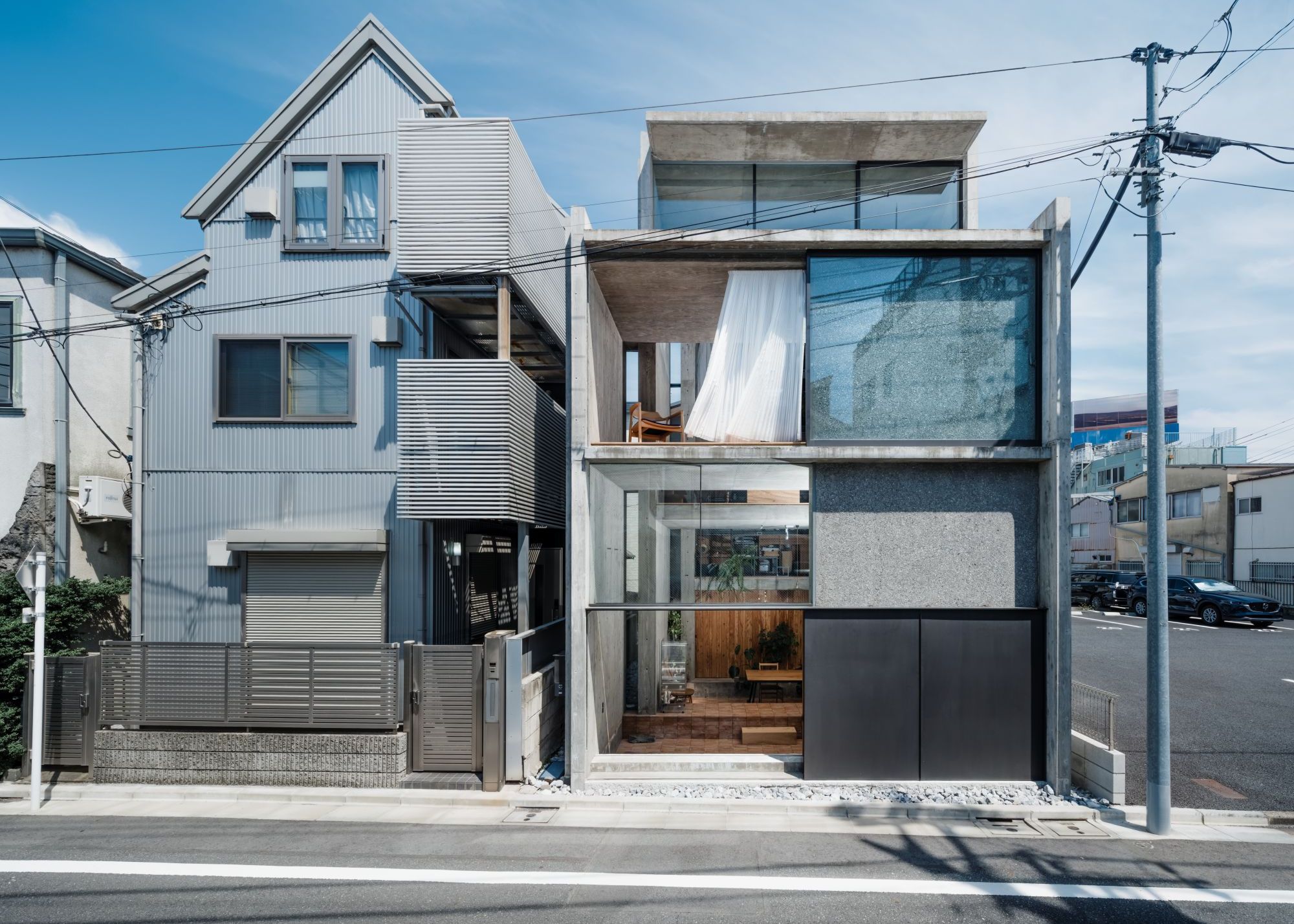 An open multi-layered Japanese home from the outside made almost entirely of concrete, next to a more closed home on a Japanese street corner on a clear blue-skied day