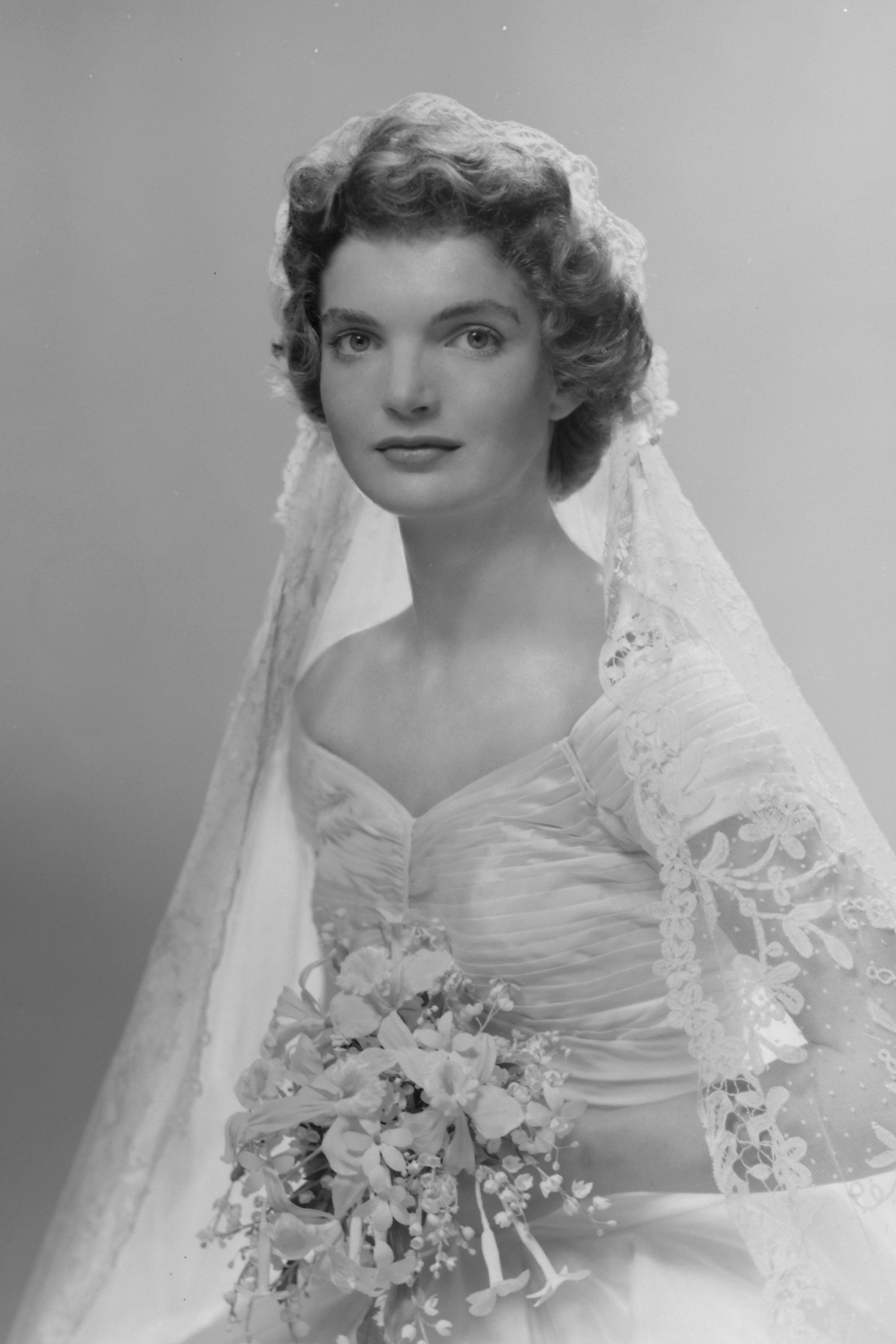 Bridal portrait of Jacqueline Lee Bouvier shows her in an Anne Lowe-designed wedding dress, a bouquet of flowers in her hands containing Lily of the Valley, Stephanotis and Orchids.