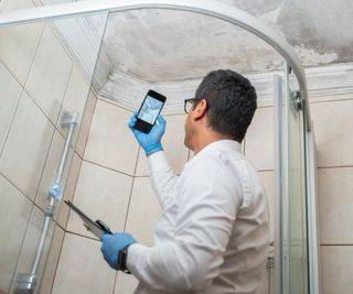 clothed man with clipboard stood in shower cubicle with phone taking picture of mould on ceiling