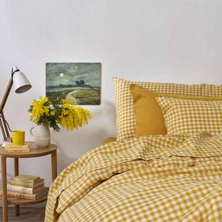 A bed dressed with yellow gingham linen bed linen and a wooden bedside table holding a lamp and a vase with yellow flowers