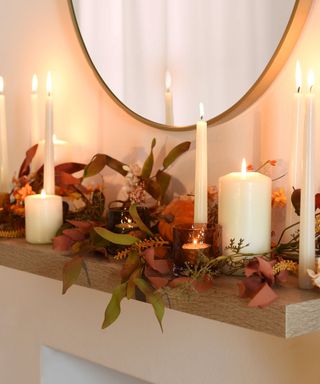 A wooden shelf with cream candles and fall leaves on top of it, with a wooden mirror above it