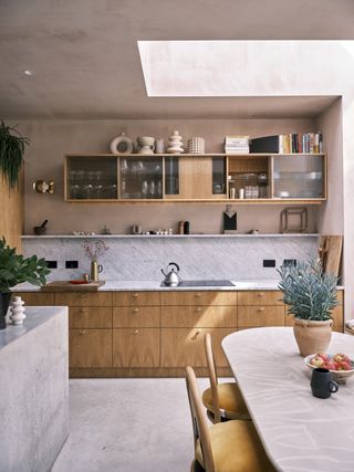 A kitchen with a concrete flour and oak fronted cabinets, shelves and a grey marbled splashback, with raw pink plaster walls
