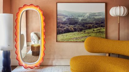 A dusty pink living room with a curved ochre yellow sofa. There is a silk pendant floor lamp in the corner and a picture of a forest hillside hanging on the wall. In the corner, there is a full-length mirror that has a futuristic sunset-colored trim.