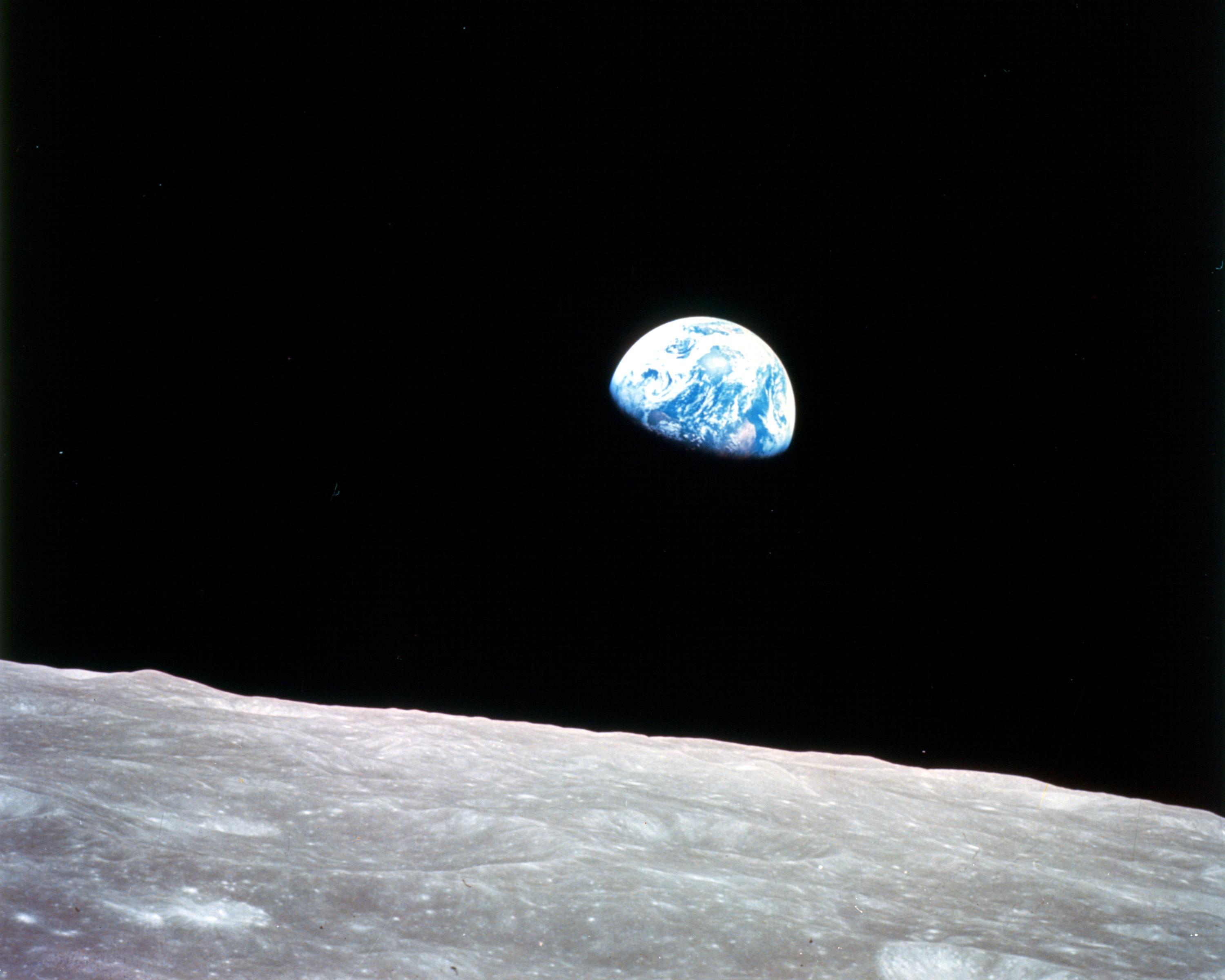 the surface of the moon is in the foreground of the image while a small blue 'marble' which is Earth appears in the background against the black backdrop of space.