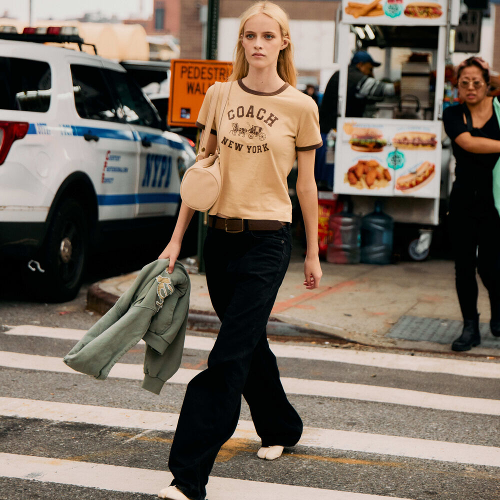 Woman walks across the crosswalk while wearing coach tee, jeans, white tabis, and a Coach kiss lock barrel bags. 