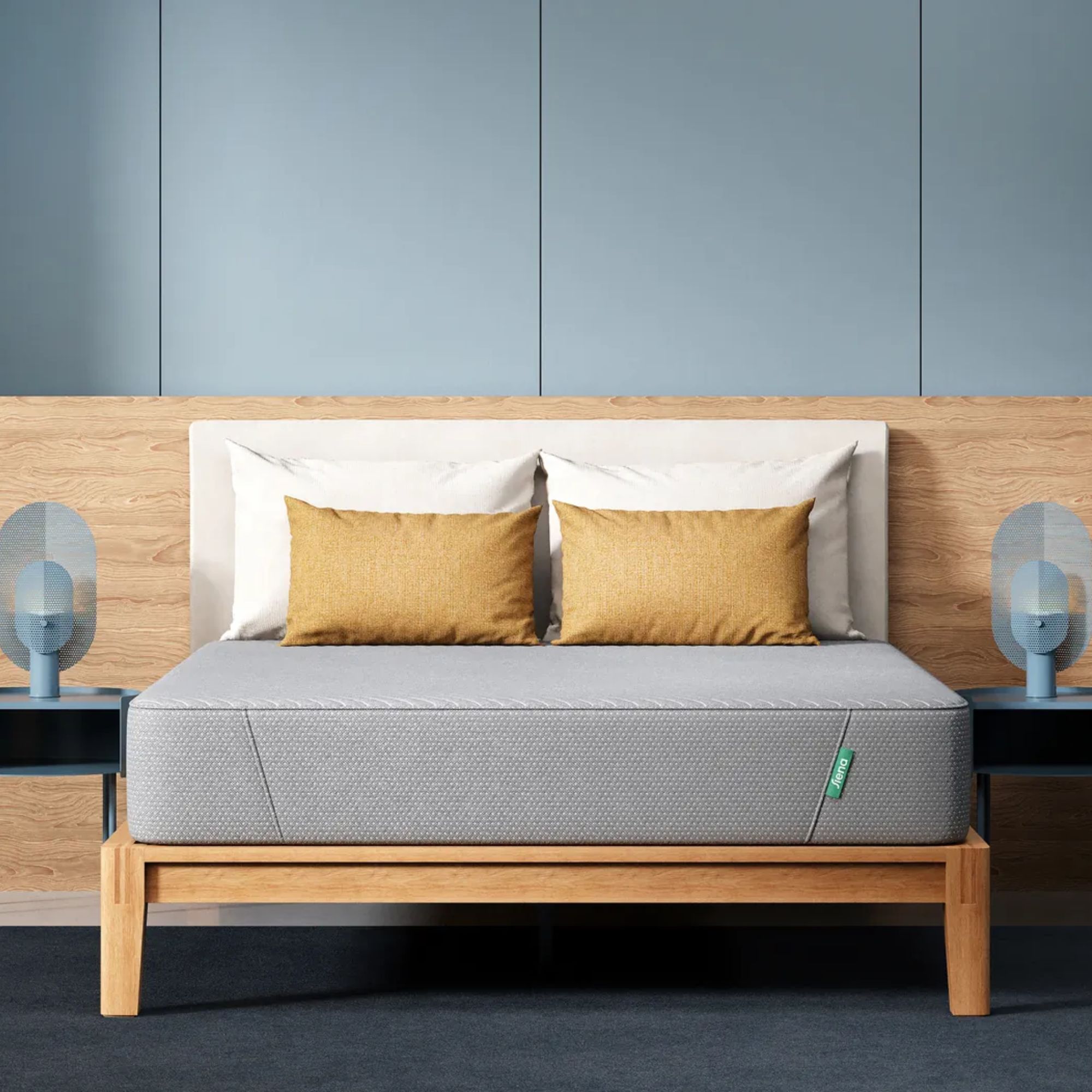 A grey Siena Signature Memory Foam Mattress on a wooden bedframe, with a white headboard, two white pillows, and two matching ochre yellow rectangular cushions. To either side of the bed are matching teal blue bedside tables with modern blue lamps. Behind the bed is a wooden wall, with blue panelling above. The floor is dark blue carpet.