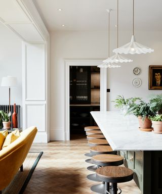 White open plan kitchen with living area, wall paneling on wall and ceiling in-between two living spaces, yellow couch on left, kitchen island on right, row of bar stools