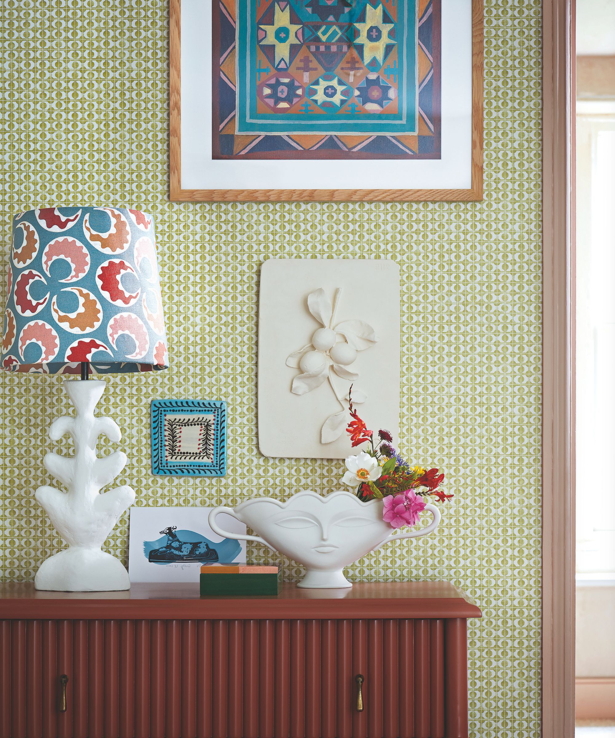 A bright and colorful green patterned wallpapered room with a brown dresser, a white abstract lamp with colorful artwork, and a white vase filled with flowers