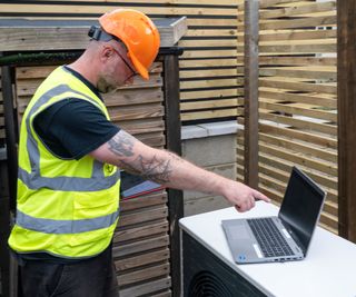 engineer wearing hi vis vest and orange hard hat pointing at laptop placed on top of air source heat pump