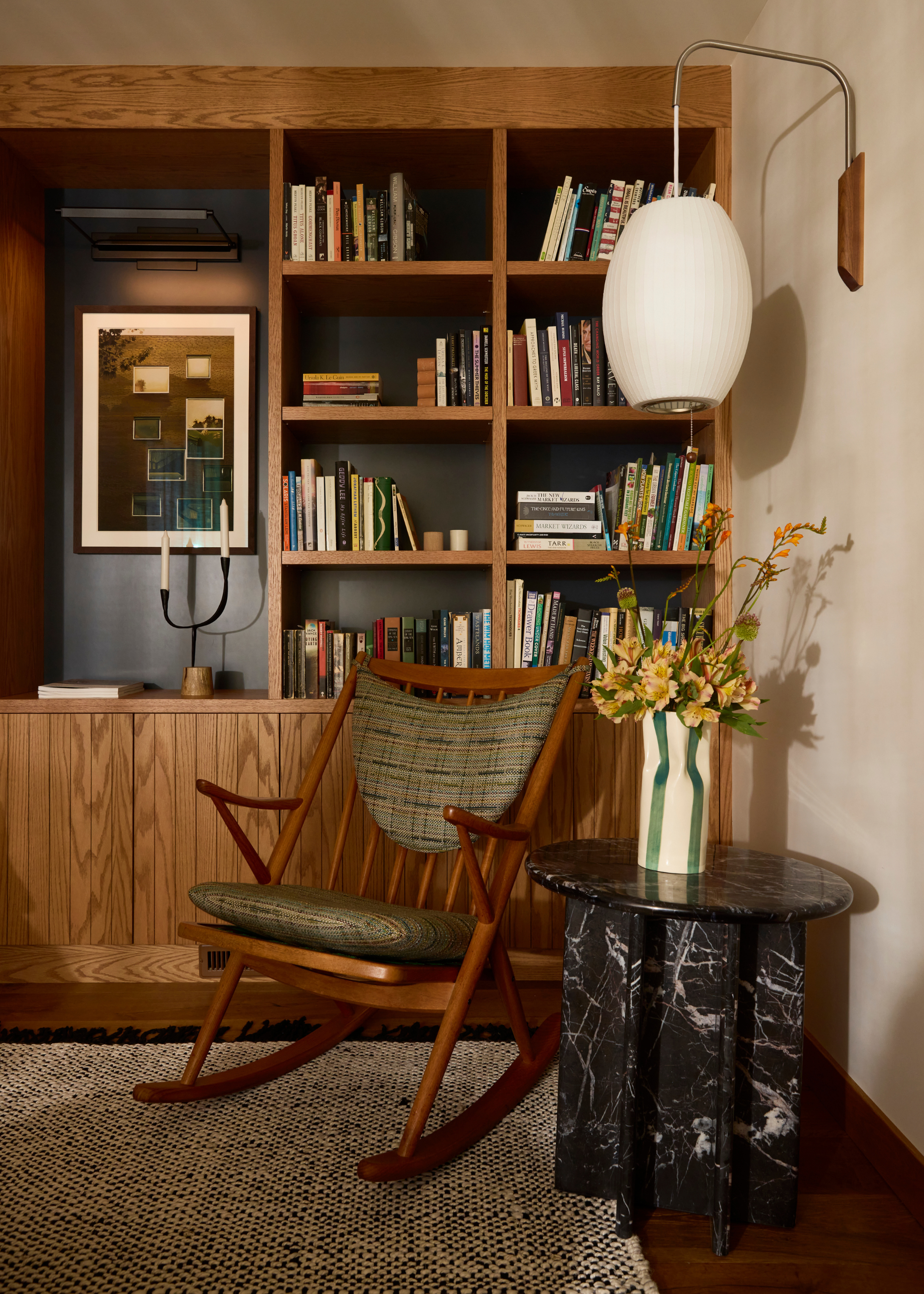 A white living room with a wood bookshelf, a wood rocking chair with green cushions, a patterned rug, and a black marble side table with a striped vase of yellow flowers underneath a hanging wall sconce
