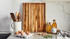 Image of a wooden cutting board leaning against a white marble backsplash on a white countertop. There is a white white utensil holder with wooden utensils in it, eggs, and a honey tray around the cutting board. 
