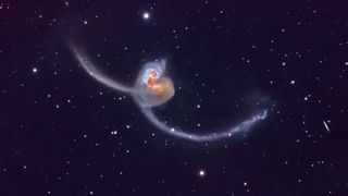 Two galaxies are pictured colliding in a dark sky dotted with stars. The orange galactic cores are visible in the center of the image and long antennae-like spiral arms can be seen stretching into space on either side.