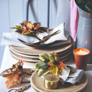 A dining table with stacks of plates and napkins decorated with autumn foliage