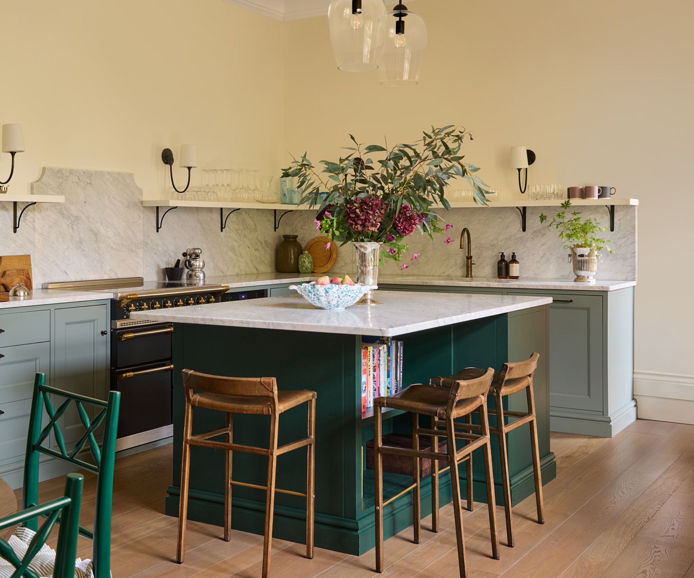 A green kitchen with light green cabinets, marble countertops, and a dark green island with a mix of closed cabinet storage and open shelves