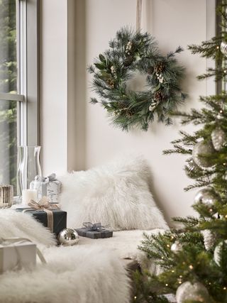 Christmas decor in window seat with Christmas wreath on the wall, tree in foreground, faux fur cushions and throws, presents, baubles, hurricane lamp