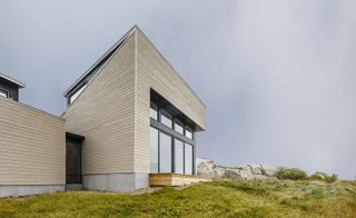 Close up exterior view of Float House during the day. The house features horizontal wood panels and tall windows in a dark coloured frame and sits on a rocky and grassy terrain