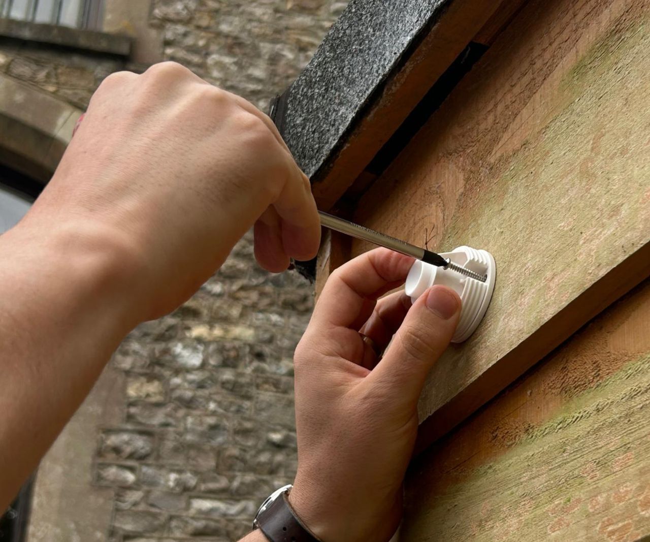Two hands screwing a white camera bracket into wooden fence post using a pen as a tool.