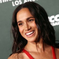 Meghan Markle wearing a red sleeveless dress and smiling in front of a step and repeat