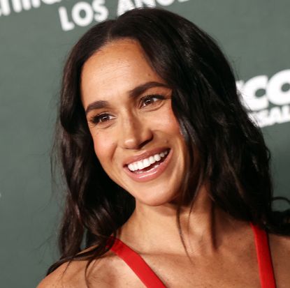 Meghan Markle wearing a red sleeveless dress and smiling in front of a step and repeat